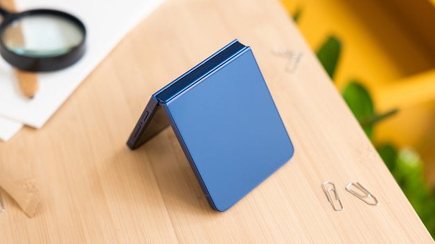 Ein Samsung Galaxy Z Flip7 Smartphone in Blau, teilweise geöffnet auf einer Holzoberfläche mit Büromaterialien in der Nähe.