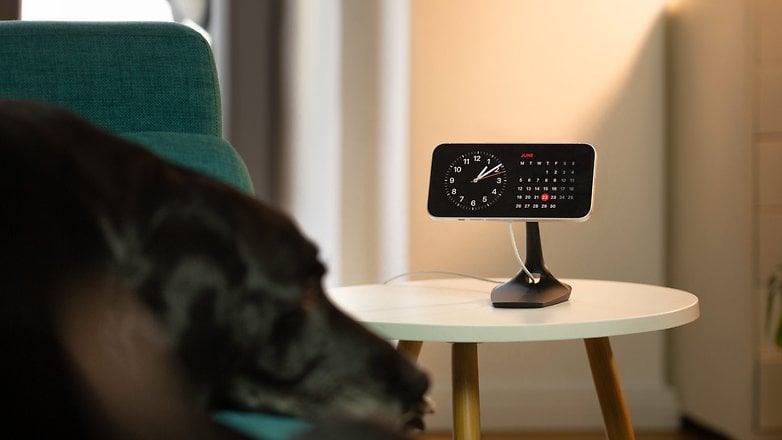 An iPhone displayed on a table, showcasing the Smart Display feature of iOS 17, while a dog lounges on the couch.