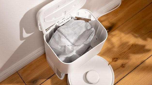 A white trash can with the lid partially open showing a gray bag inside.