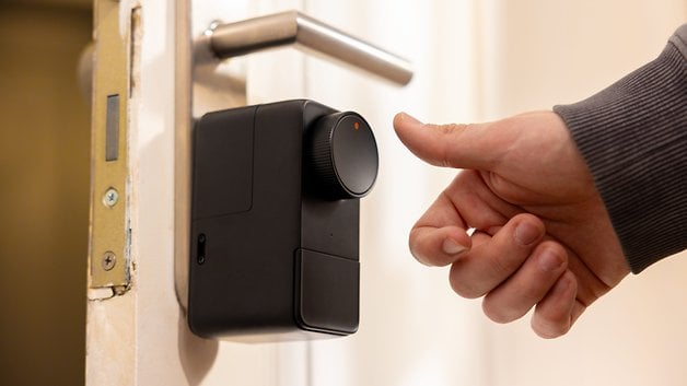 A hand uses a SwitchBot Lock Pro on a door, shown with a button and a slight red light.