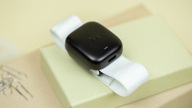 A black wearable device on a white strap, resting on a light green box with paper clips nearby.