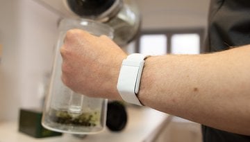 Una mano sostiene una tetera de vidrio con un reloj inteligente blanco en la muñeca.