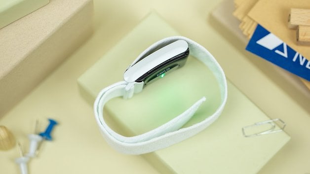 A white wearable device with a green light, resting on a light surface with various office supplies around.