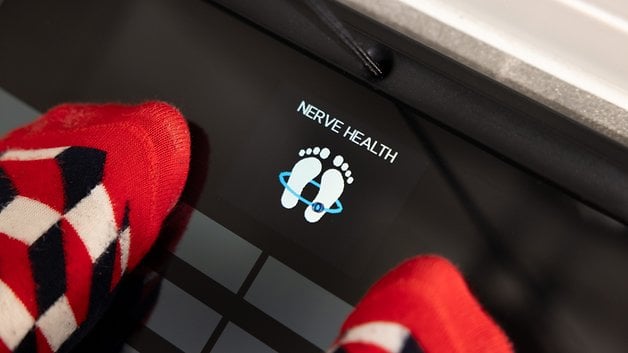 A person's feet in colorful socks on a Withings body scan device displaying 'NERVE HEALTH'.