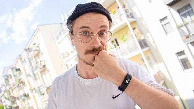 A person with glasses and a mustache holds their chin, wearing a Garmin Vivosmart 5 smartwatch.