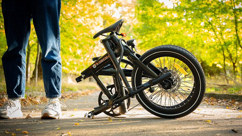 Ein gefaltetes schwarzes Fahrrad steht neben einer Person in Jeans und Sneakers in einem Park.