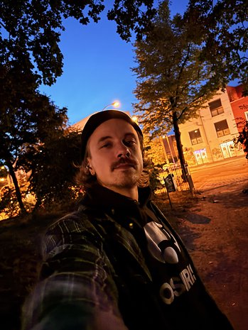 Uma pessoa tirando uma selfie em um parque ao anoitecer, com árvores e postes de luz ao fundo.