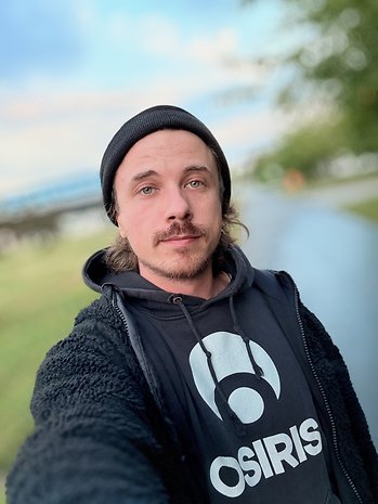 Um homem com bigode usando um gorro preto e um moletom ao ar livre.