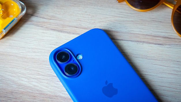 Ein blauer Apple iPhone 16 mit zwei Kameras auf einer Holzoberfläche, daneben gelbe Kopfhörer und Sonnenbrille.