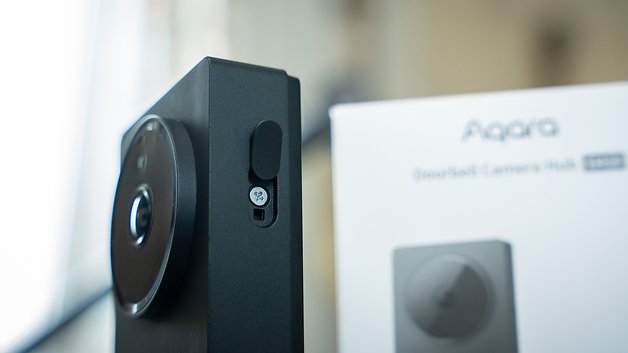 Aqara Türklingel-Kamera-Hub mit kreisförmigem Objektiv und seitlichem Knopf, neben der Verpackung.