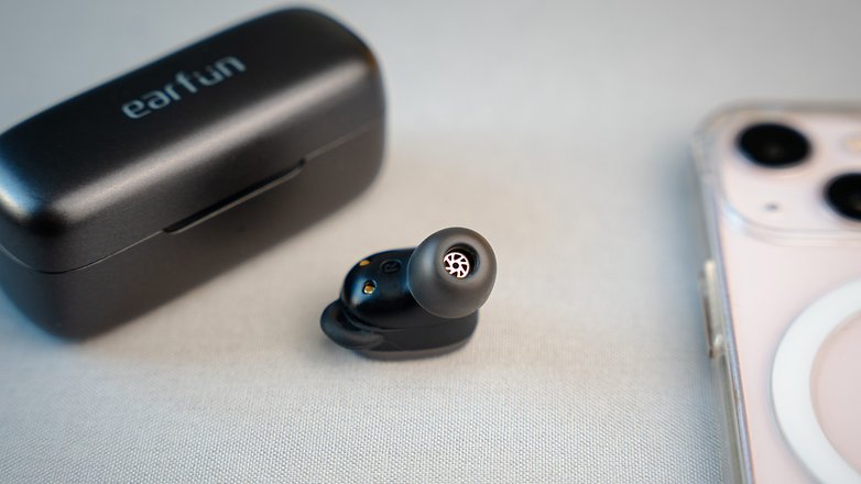 Un des écouteurs EarFun Free Pro 3 posé entre un smartphone à droite et son boîtier de recharge à gauche