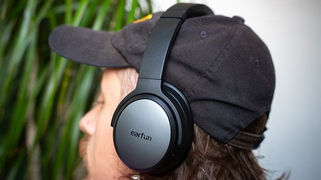 A man with long hair wearing a black cap and EarFun Tune Pro headphones.