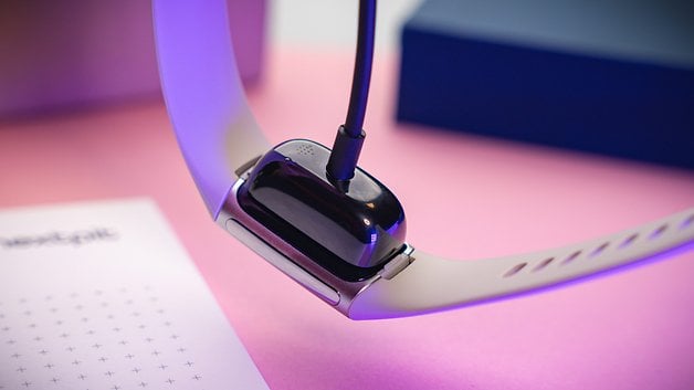 Fitbit Charge 6 charging on a pink surface, with a charger connected.