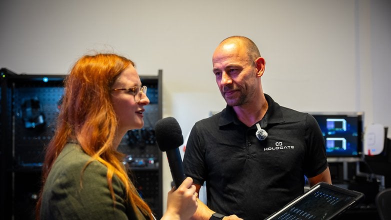 Une femme interviewe un homme tenant une tablette, avec un holodeck en arrière-plan.