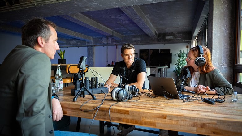 Three people discuss in an office setting with microphones and headphones around a wooden table.