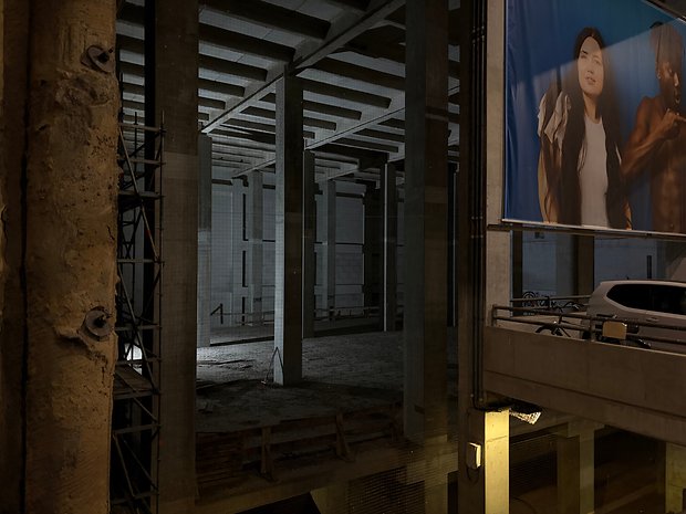 Eine leere Baustelle, beleuchtet von einer einzelnen Lichtquelle, mit Betonsäulen und einem großen Werbebanner.