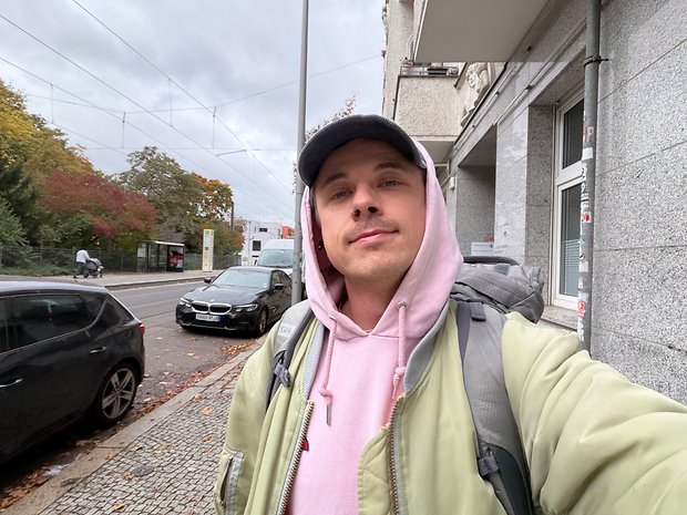 Eine Person steht auf einer Straße mit einem Rucksack, trägt einen rosa Hoodie und eine grüne Jacke, mit Autos und Bäumen im Hintergrund.