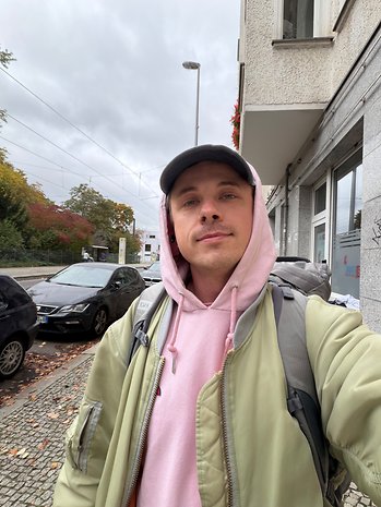 Ein junger Mann in einer grünen Jacke und rosa Hoodie steht auf einer Stadtstraße bei bewölktem Himmel.