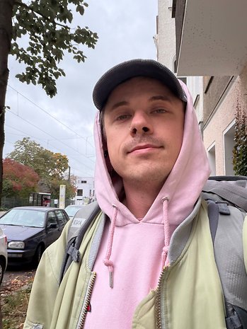 Eine Person trägt einen rosa Hoodie und eine grüne Jacke und steht im Freien mit Autos und Bäumen im Hintergrund.