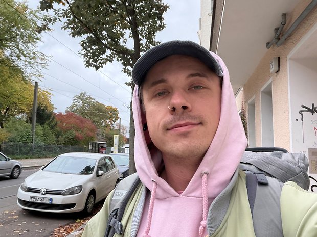 Eine Person mit einem rosa Hoodie und einer Mütze steht auf einer Straße mit Bäumen und geparkten Autos.