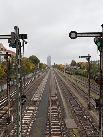 Ein Schienenweg, der sich unter einem bewölkten Himmel in die Ferne erstreckt, mit grünen Signalen und Stadtgebäuden im Hintergrund.