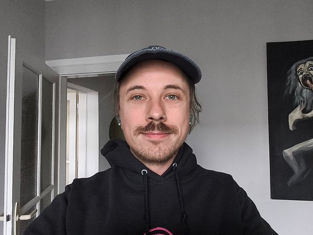 Ein junger Mann mit Schnurrbart und Cap, der in einem Raum mit einem Gemälde an der Wand lächelt.