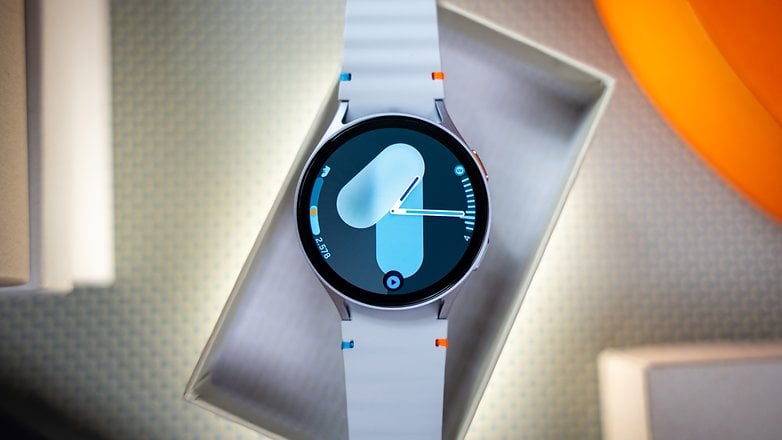 A Samsung Galaxy Watch 7 displayed in a box with a blue and white strap showing a digital watch face.