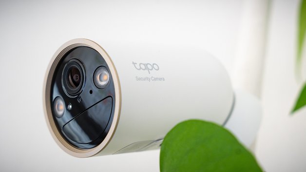 Tapo C460 Sicherheitskamera mit zwei Linsen und LED-Leuchten, teilweise von einem grünen Blatt verdeckt.