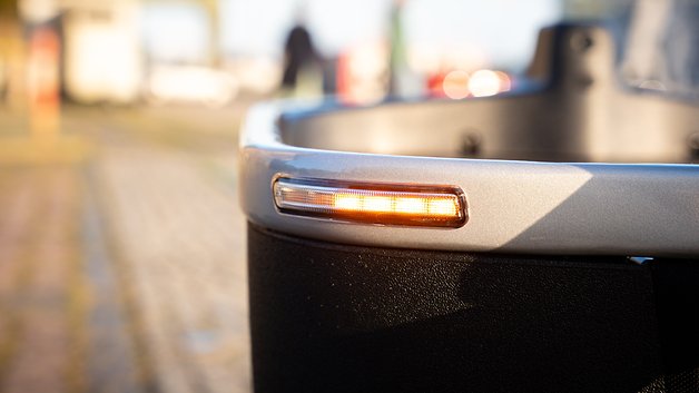 Nahaufnahme eines silbernen Fahrzeugteils mit leuchtendem bernsteinfarbenem Licht, vor verschwommenem Hintergrund.