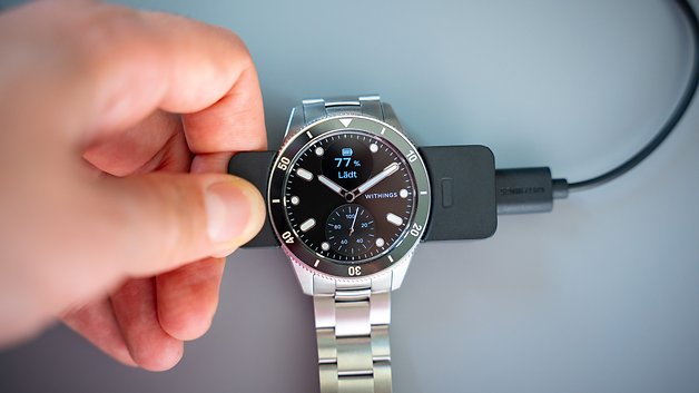 Eine Hand hält eine Withings ScanWatch Nova, während sie auf einer Dockingstation aufgeladen wird, zeigt 77% Akku an.