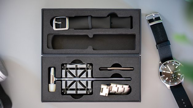 Offene Box mit einem schwarzen Uhrenarmband, Werkzeugen und einer silbernen Uhr.