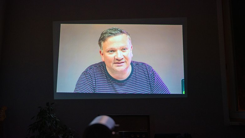 Un appel vidéo projeté sur un mur montrant un homme avec des cheveux courts portant un t-shirt rayé.