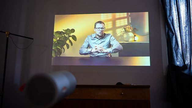Ein Projektor zeigt einen Mann in einem Hemd, der vor einem bunten Hintergrund spricht, mit einer Pflanze und einer Lampe im Blick.