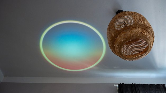 Eine farbenfrohe kreisförmige Lichtprojektion an der Decke neben einer gewebten Pendelleuchte.