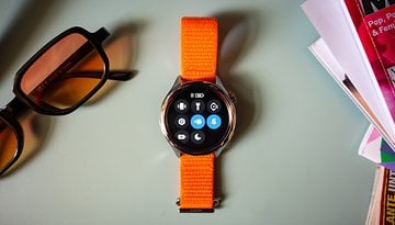 Une montre Xiaomi S4 avec un bracelet orange, affichant diverses icônes d'applications, à côté de lunettes de soleil et de magazines.