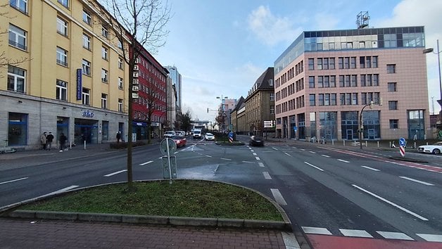 Vue de rue avec des bâtiments, des arbres et des voitures sous un ciel partiellement nuageux.