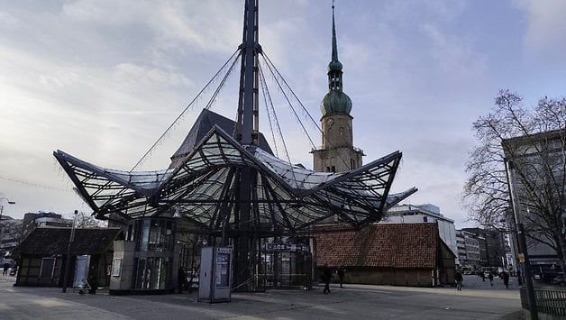 Eine moderne Glasstruktur, die einer Blume ähnelt, mit einem Kirchturm im Hintergrund.