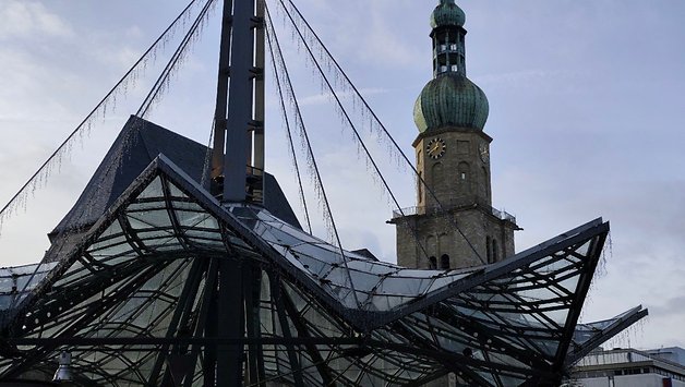 Une structure en verre moderne avec un toit pointu au premier plan et une tour de l'horloge à l'arrière-plan.