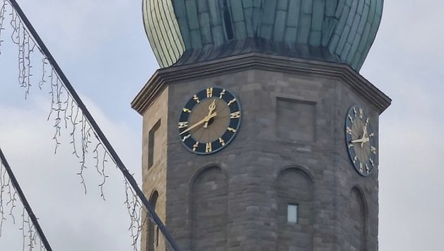 Nahaufnahme eines Uhrturms mit drei Zifferblättern und einer grünen Kuppel unter einem bewölkten Himmel.