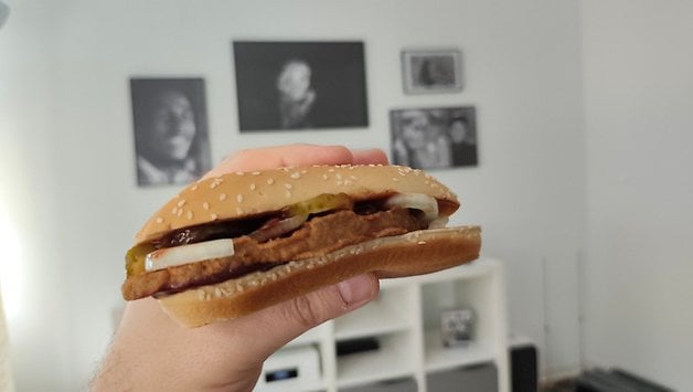 Eine Person hält ein Sesambrötchen mit einem Fleischpatty, Gurken und Zwiebeln in einem Innenraum.