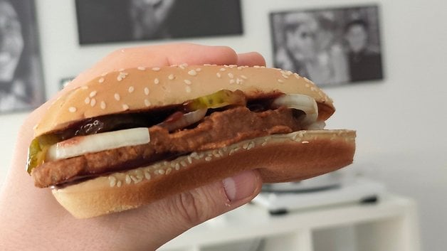 Eine Hand hält einen Sesambrötchen-Burger mit Gurken, Zwiebeln und Ketchup vor einem verschwommenen Hintergrund.