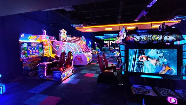 Une zone d'arcade avec des machines colorées et des lumières néon, présentant un jeu de tir 'Nerf' et un jeu de combat.