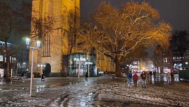 Scène nocturne d'une place avec des personnes marchant, une tour d'église et des arbres illuminés.