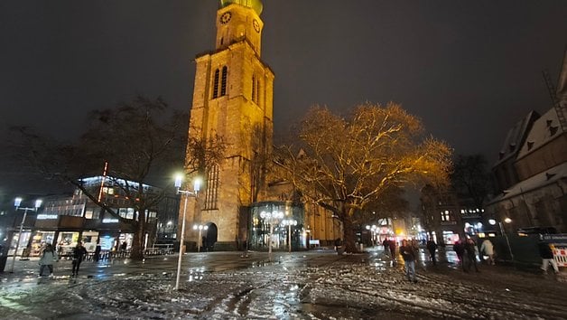 Ein historisches Gebäude mit einem hohen Uhrturm, das nachts beleuchtet ist, umgeben von Bäumen und Menschen auf einem verschneiten Platz.