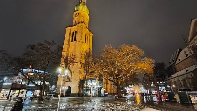 Nachtansicht eines hohen Uhrturms mit einem beleuchteten Baum und Menschen, die auf einem verschneiten Platz gehen.