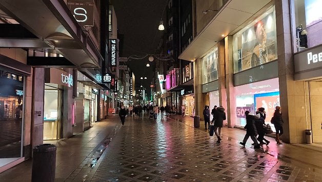 Eine belebte Einkaufsstraße bei Nacht, mit beleuchteten Schaufenstern und Passanten.