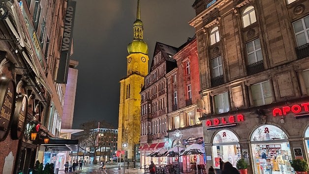 Eine nächtliche Ansicht einer Straße mit historischen Gebäuden und einem Uhrturm, unter bewölktem Himmel beleuchtet.