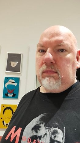 Une personne avec la tête chauve et une barbe, portant un t-shirt noir, pose devant des œuvres d'art encadrées colorées.