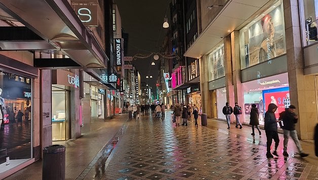 Eine belebte Straße bei Nacht mit Geschäften und Fußgängern, die Lichter auf dem nassen Gehweg reflektieren.