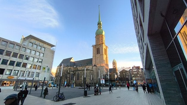 Eine historische Kirche mit grünem Turm steht auf einem belebten Platz mit Passanten.